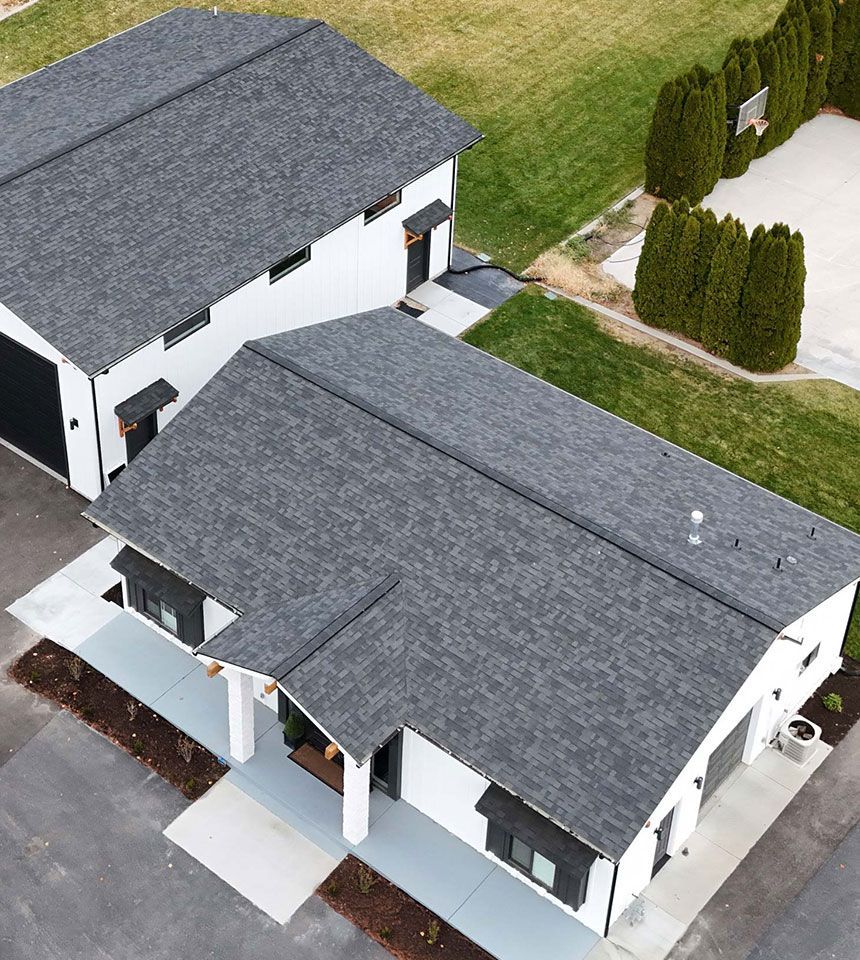 Two-story modern home and detached garage with dark gray roofs and white siding. Green grass surrounds them, with a basketball court visible nearby.