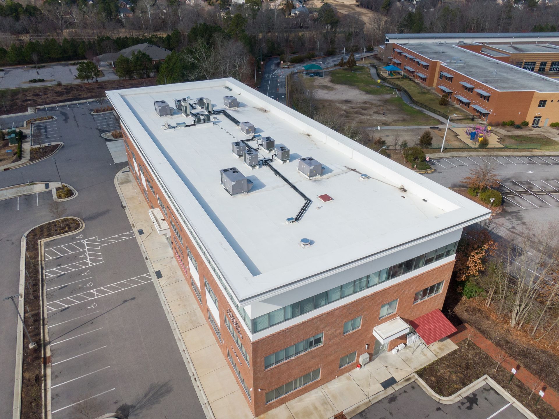 Aerial view of a rectangular brick building with a flat white roof. The building has air conditioning units on top and is surrounded by a parking lot and other structures.