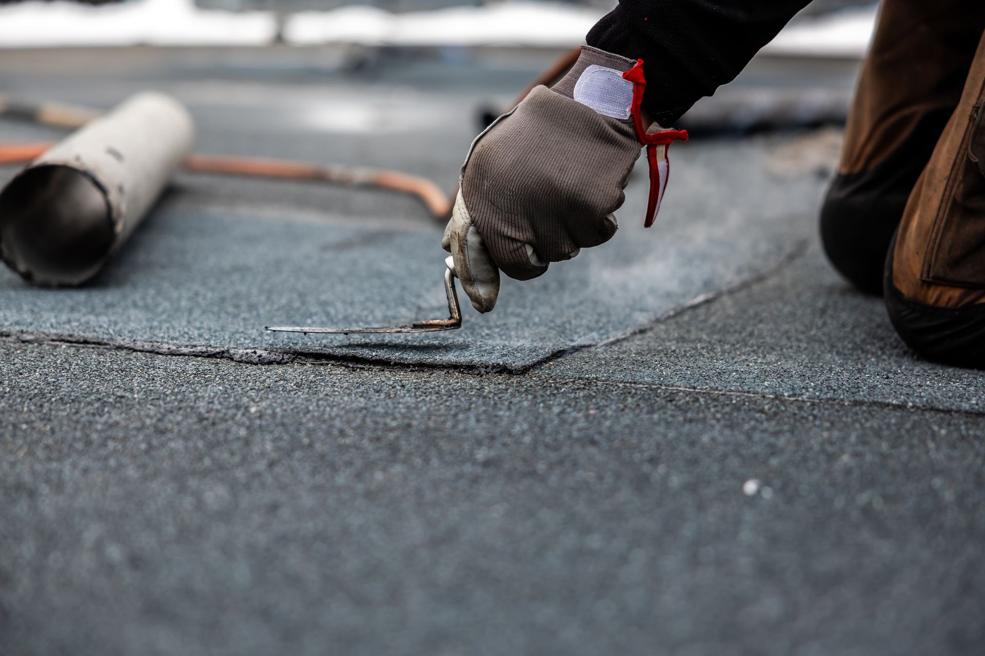 Person kneeling on a dark roof, using a tool to repair the surface; gray gloves, brown boots, and a metal pipe are visible.