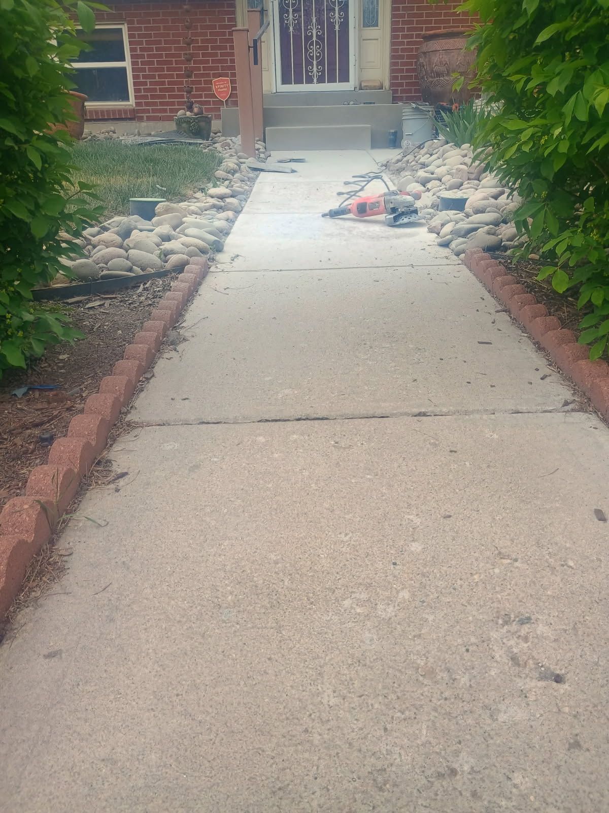 Concrete walkway leading to a front door, bordered by rocks and bushes with debris on the path