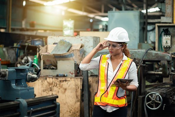 A woman in a hard hat and safety vest is using a tablet in a factory.