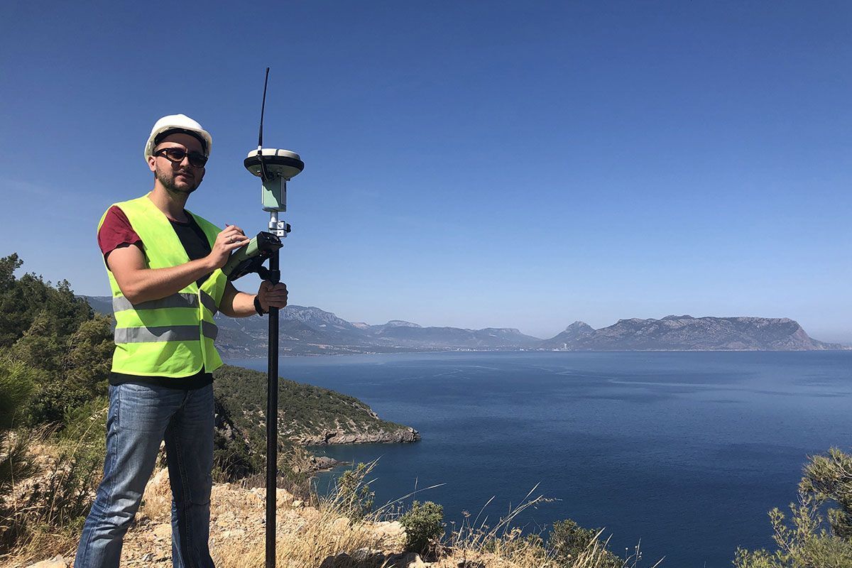 A man in a hard hat and safety vest is standing on a hill overlooking the ocean.