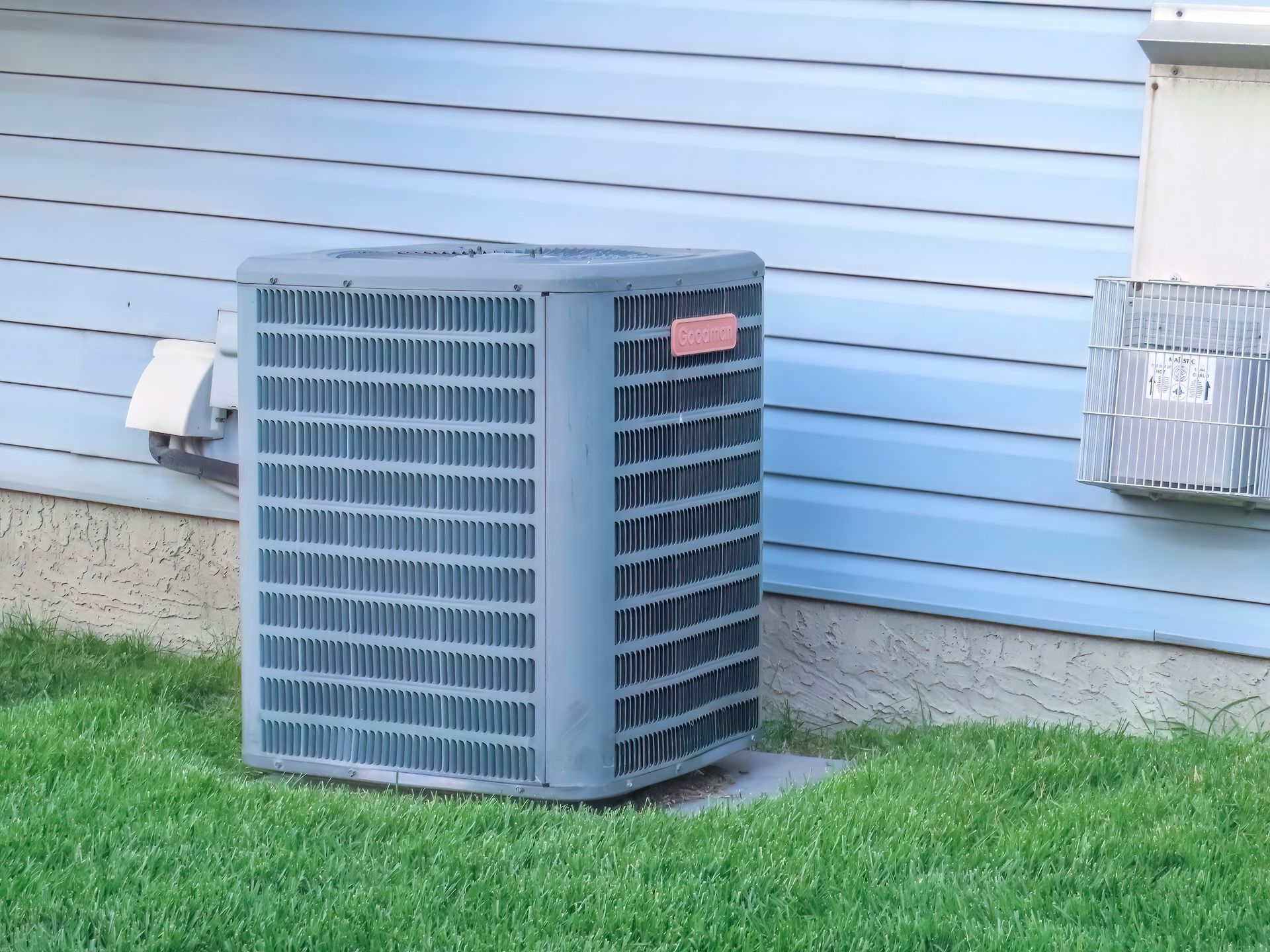 A gray central air conditioning unit sits on a concrete pad next to light blue house siding and green grass.