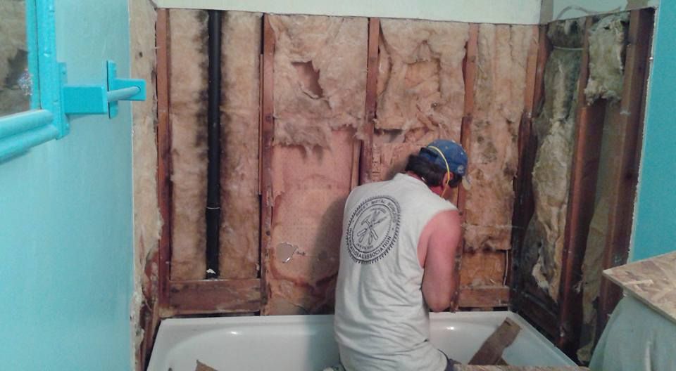 Man in a bathroom removing wall; visible insulation, black pipe, and bathtub. Blue walls.