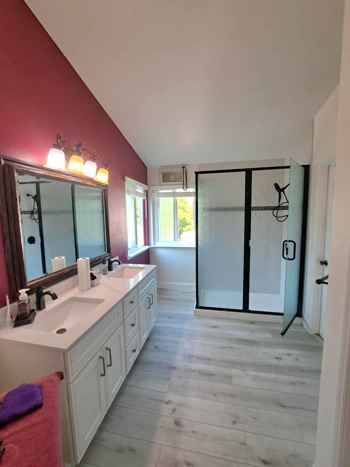 Bathroom with white vanity, large mirror, glass shower, and gray wood-look flooring. Walls are white and red.