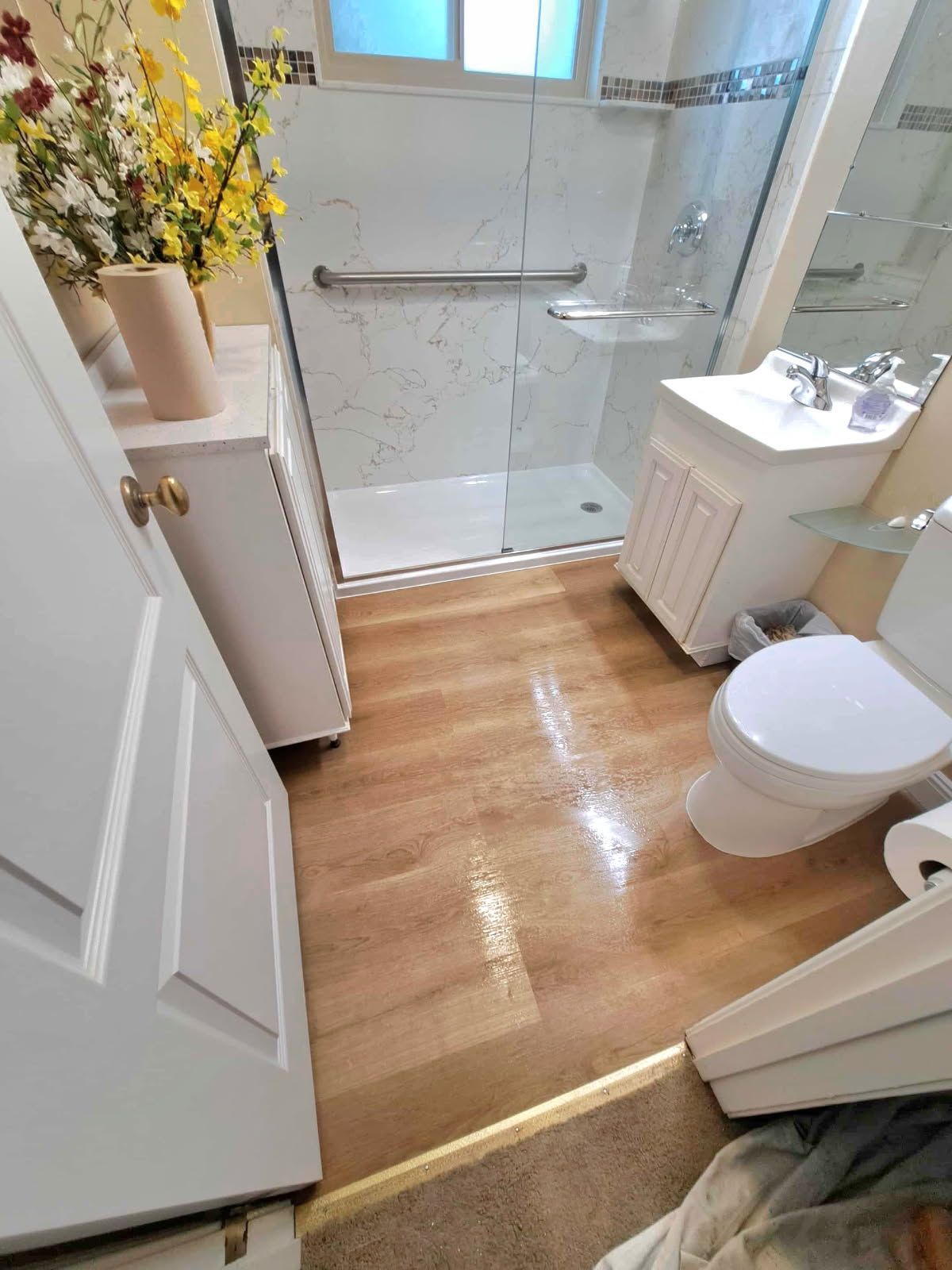Bathroom with white cabinets, shower, and toilet; wood-look floor with water spots.
