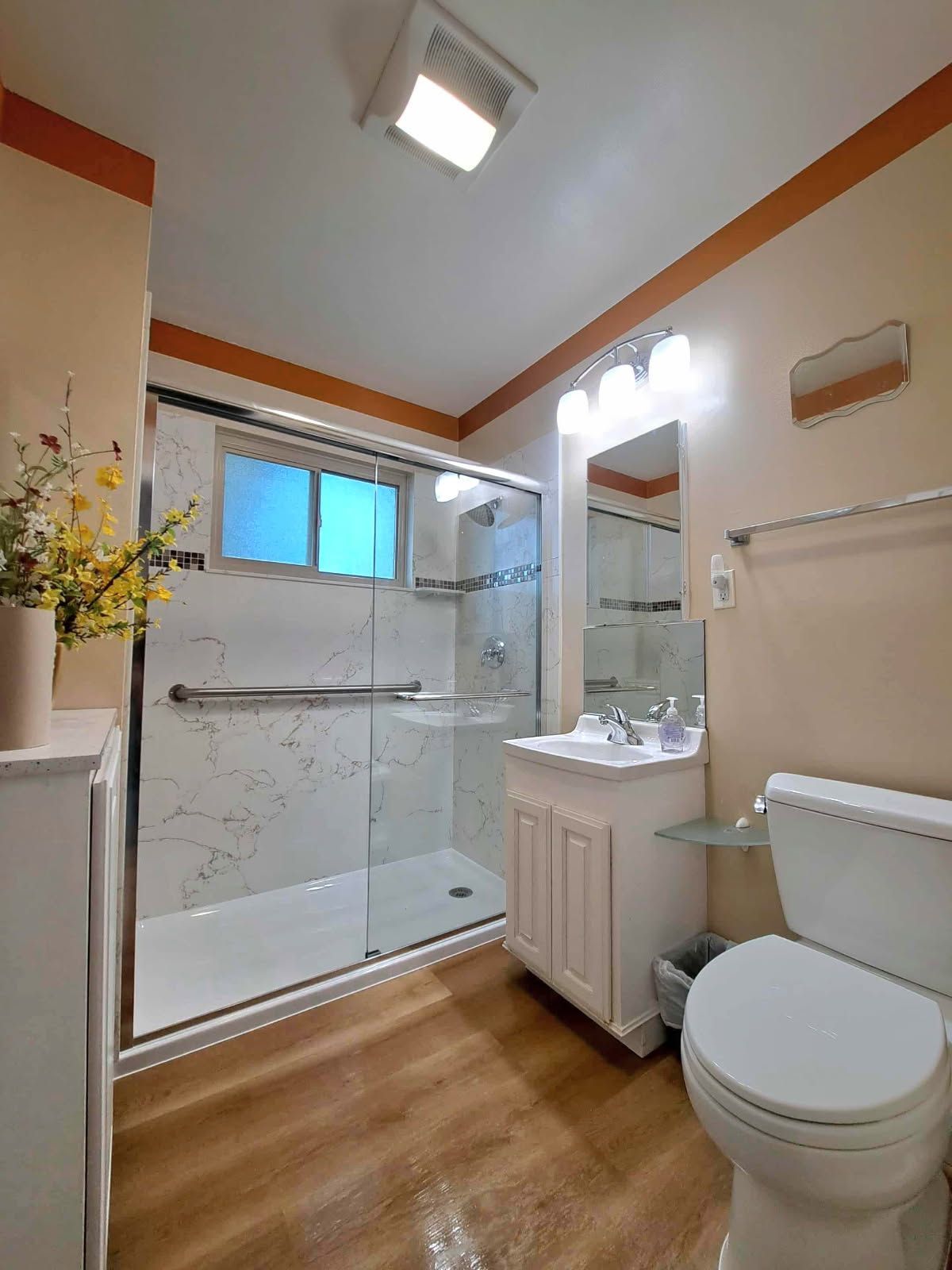Bathroom with a glass shower, toilet, and vanity on wood-look flooring. Light walls with orange trim.