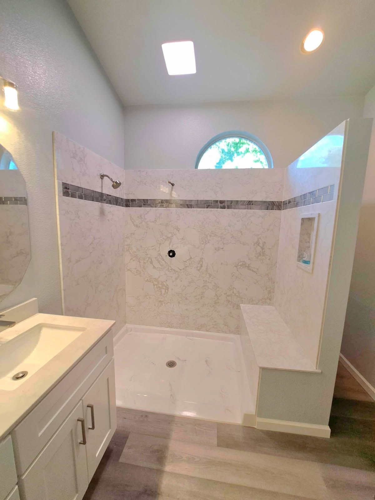 Bathroom with white tile shower, built-in bench, arched window, and vanity with a round mirror.