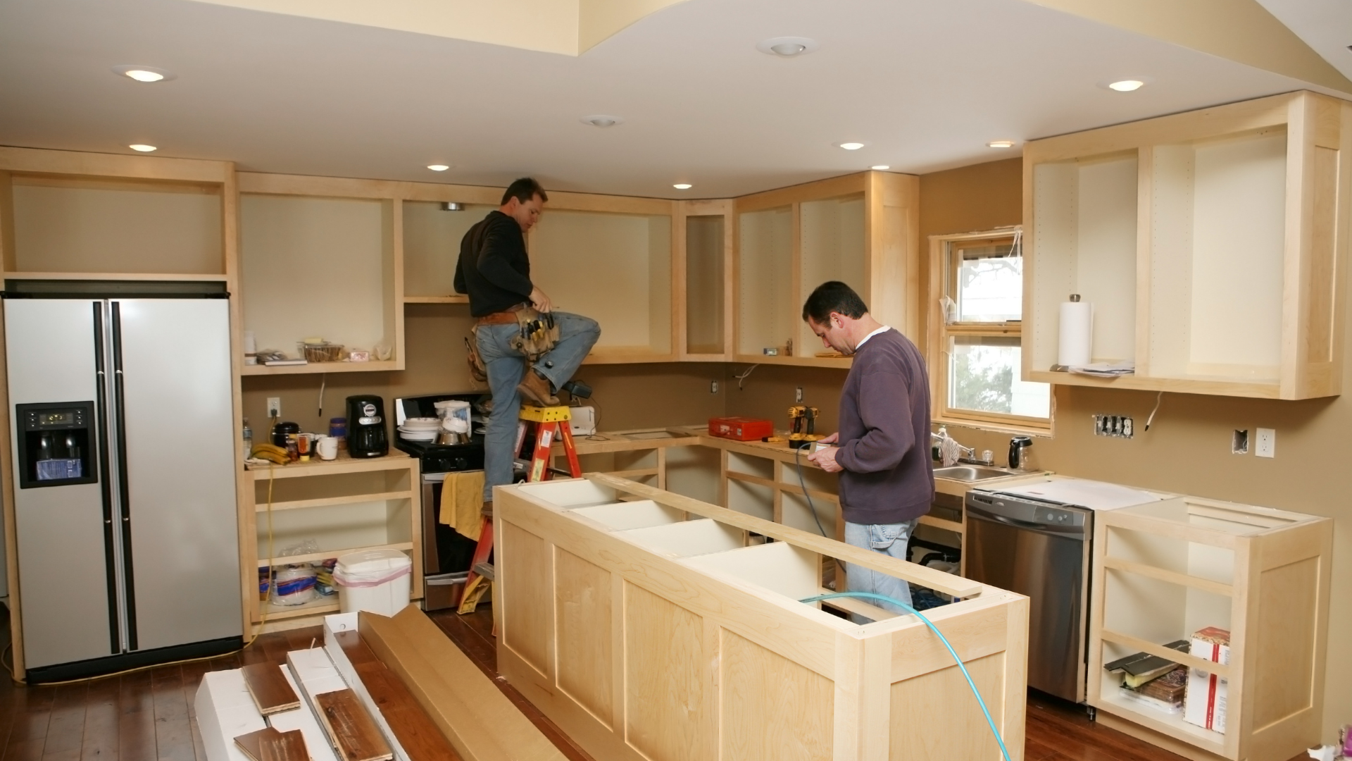Two men install kitchen cabinets in a room under renovation; the cabinets and appliances are partially installed.