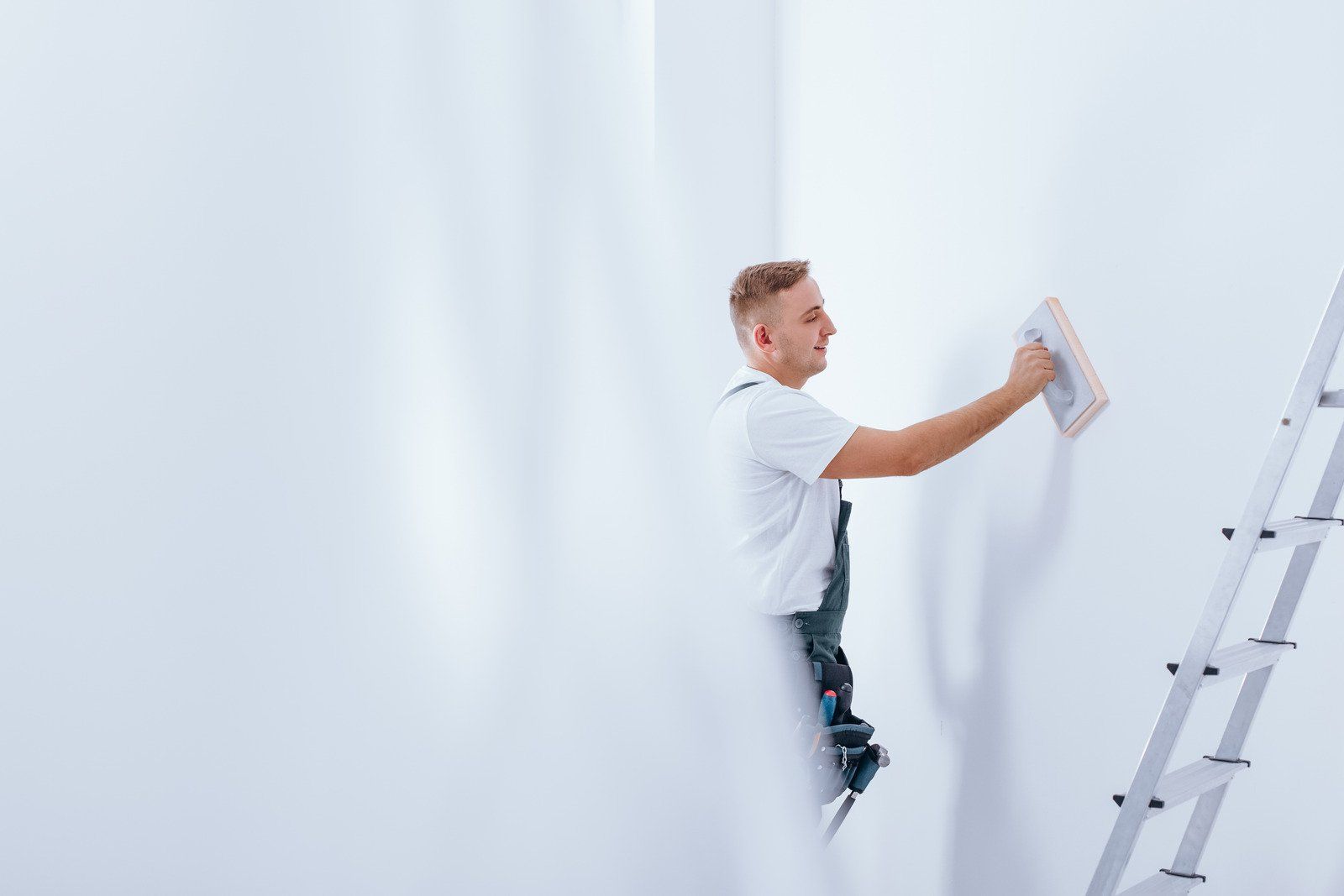Man uses a trowel to apply plaster to a white wall, leaning on a ladder.