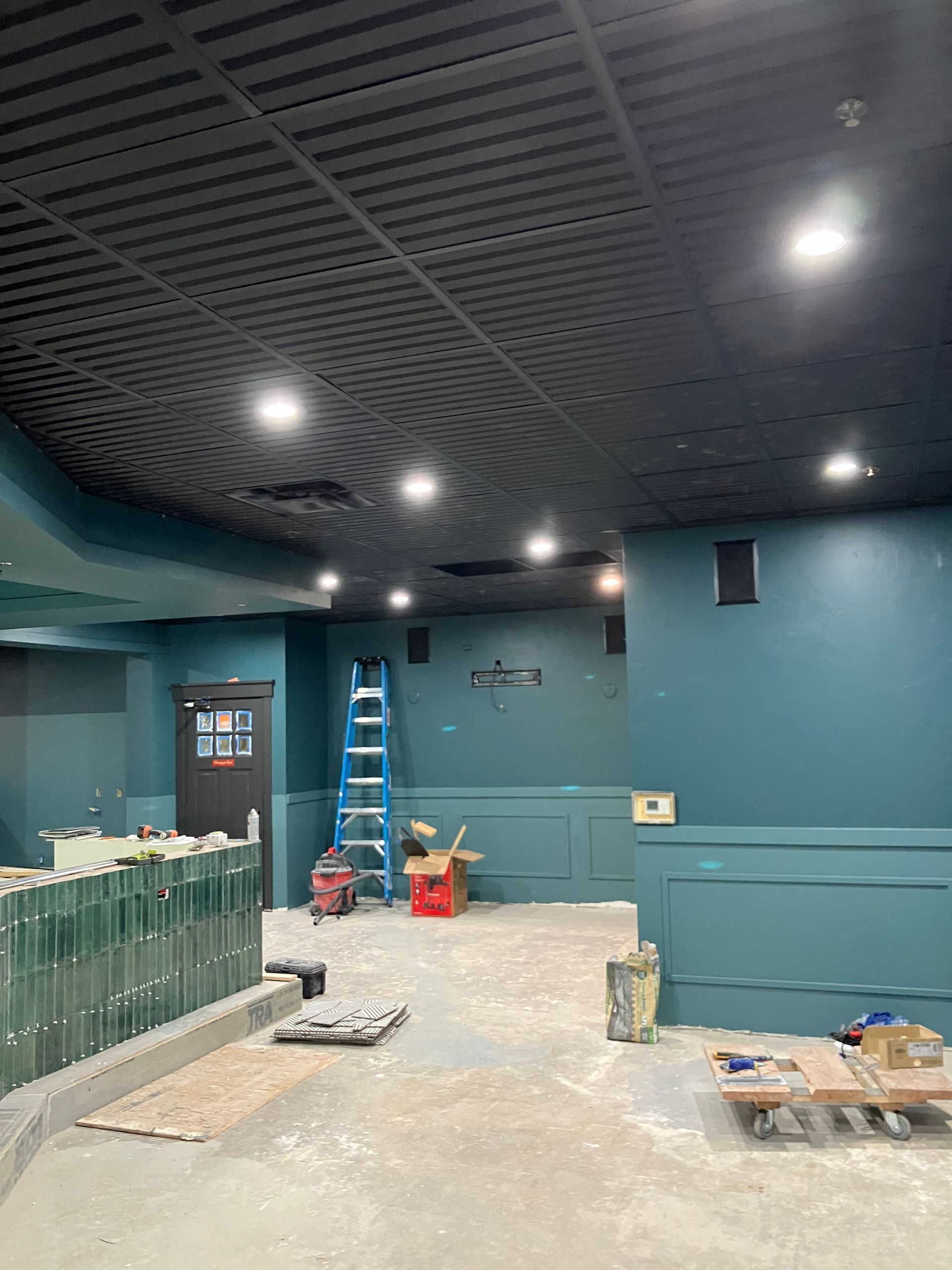 Interior under construction, teal walls, black ceiling with lights, ladder, and construction materials.