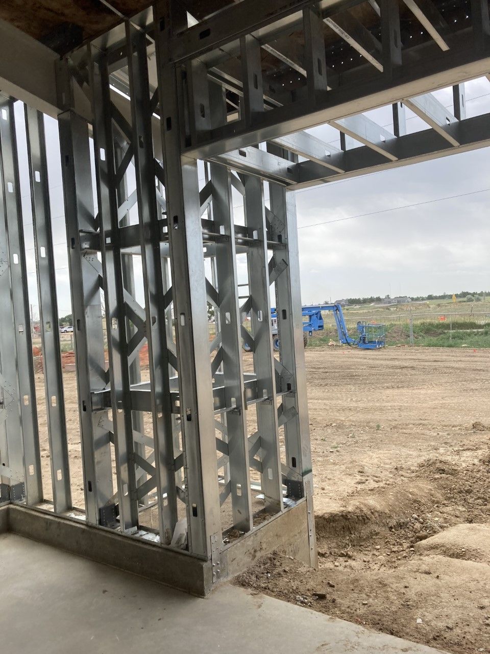 Metal framing of a building's exterior wall and doorway. Construction site with dirt and distant blue machinery visible.