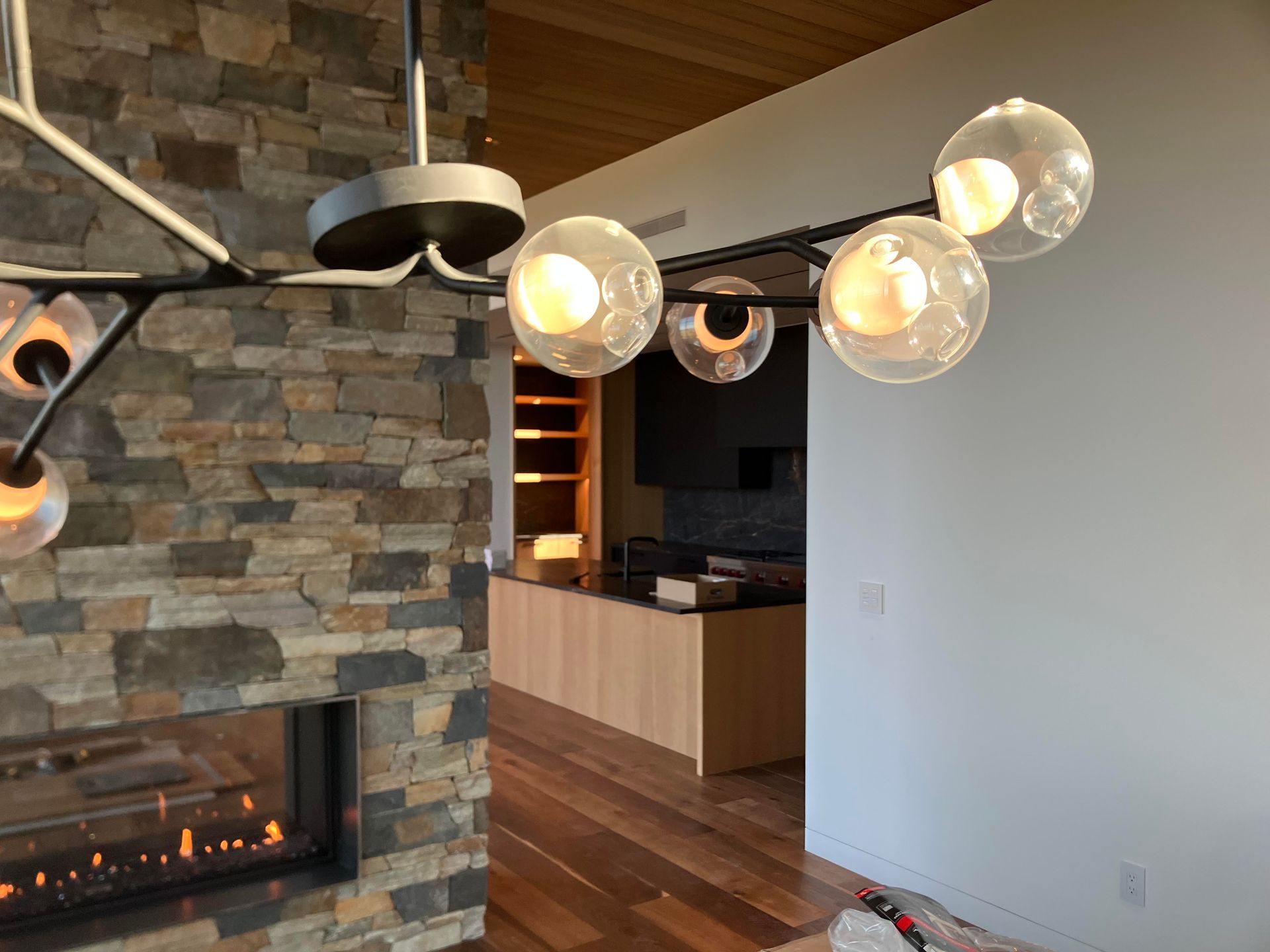 Modern chandelier with glass globe lights in a room with a stone fireplace and wooden flooring.