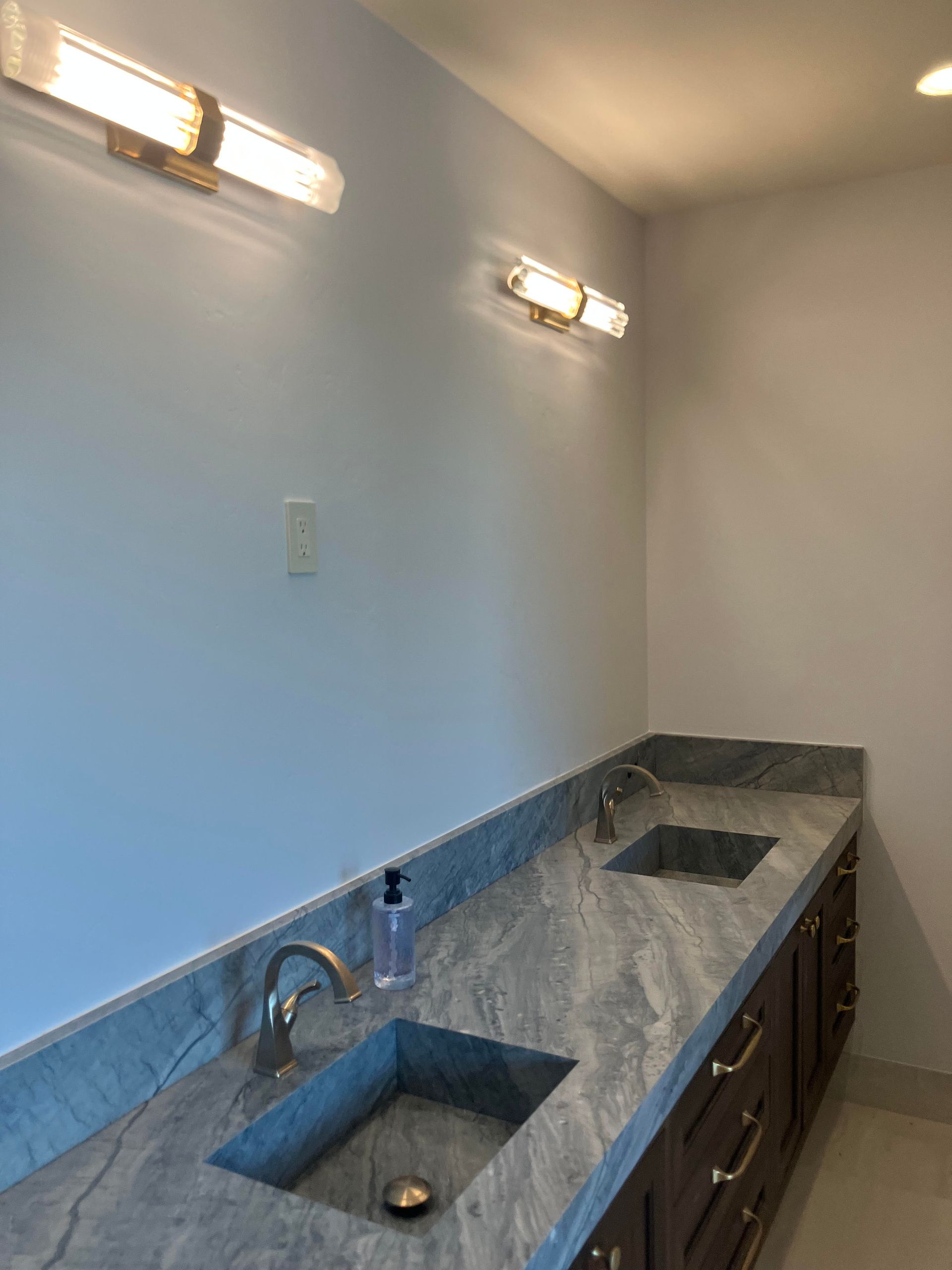 Bathroom with dual sinks and gray marble countertop, brown cabinets, and two sconces on a light blue wall.