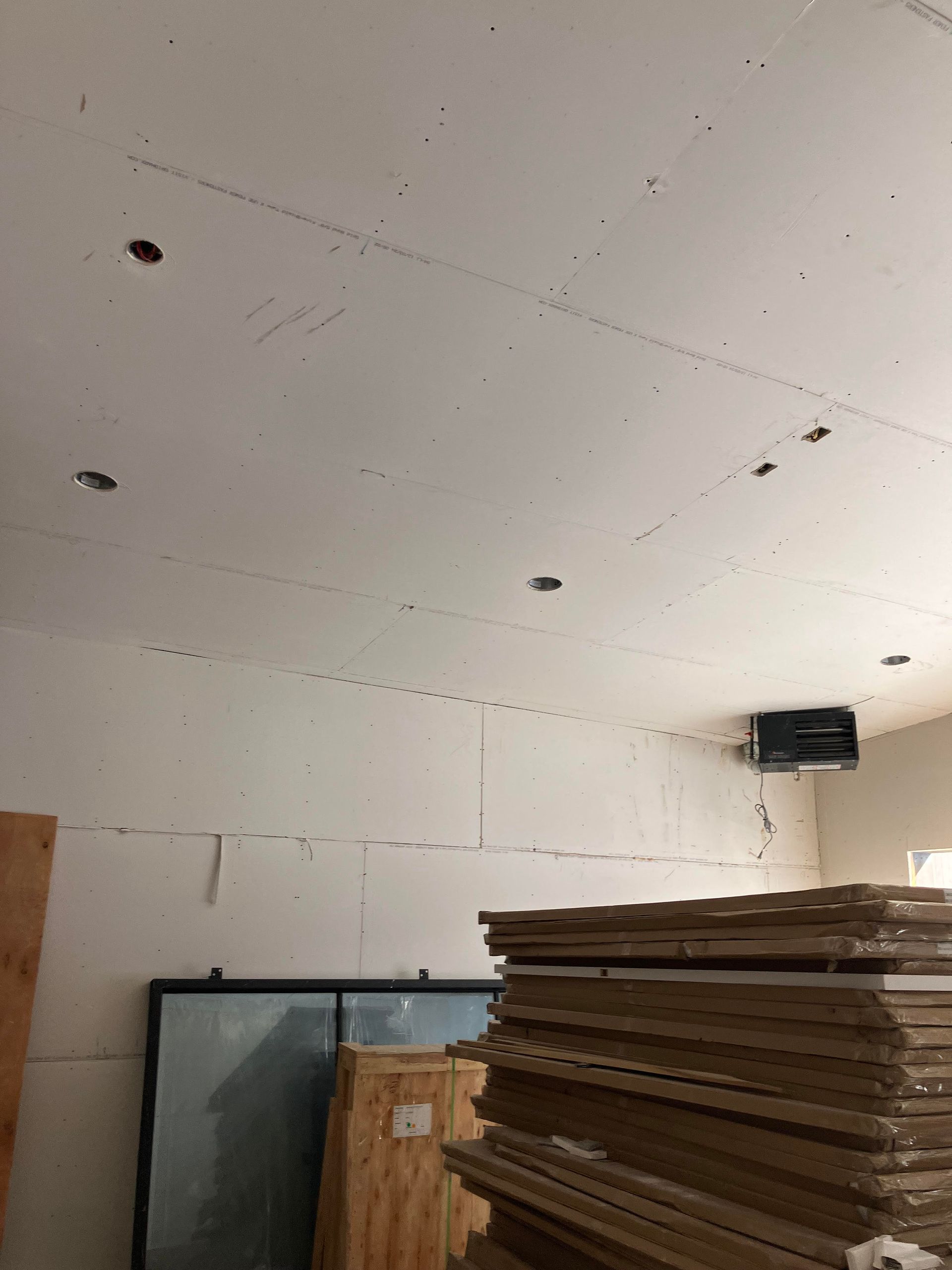 Interior of a room under construction with drywall and recessed light holes. A stack of boards is in the foreground.