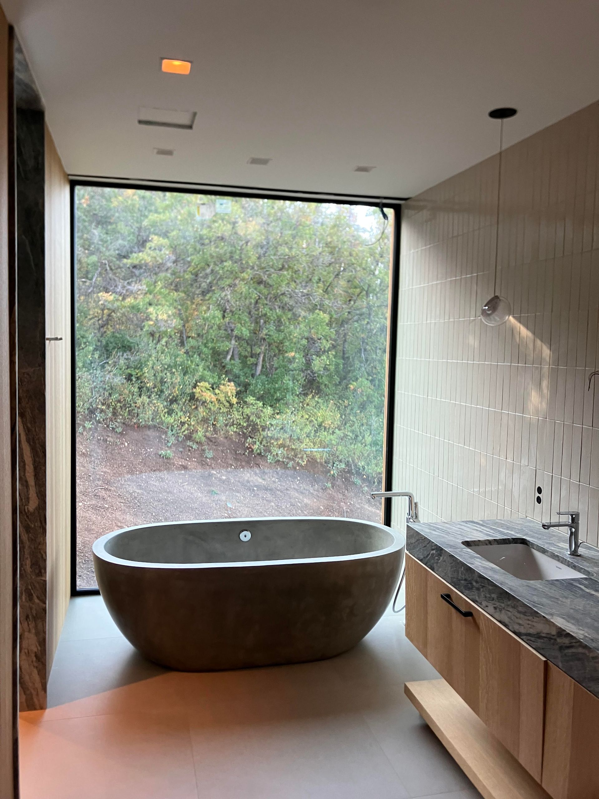 Bathroom with large window overlooking trees, featuring a stone bathtub, floating vanity, and light-colored walls.