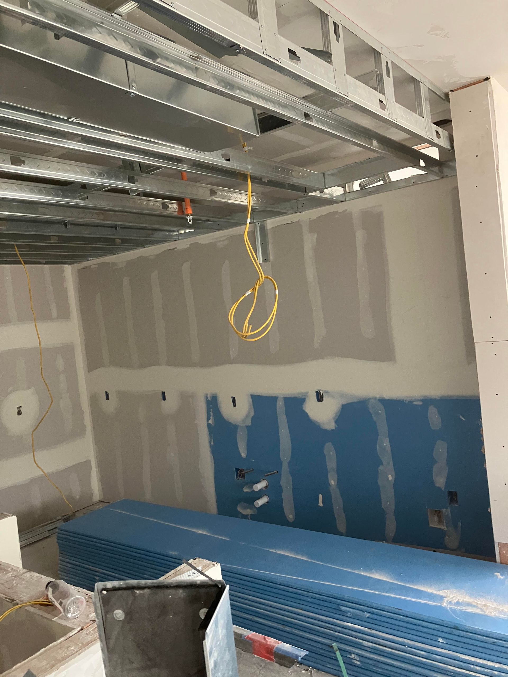Interior construction site with metal framing, drywall, and stacked blue boards. Yellow electrical cord hangs.