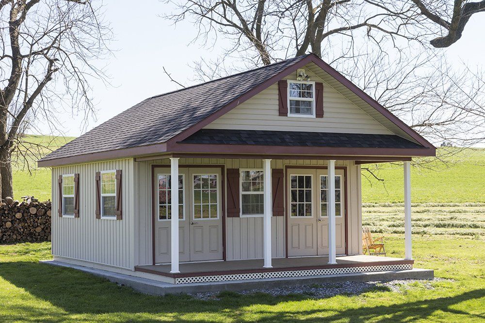 Tan and brown shed with a porch and gabled roof, set in a grassy yard.