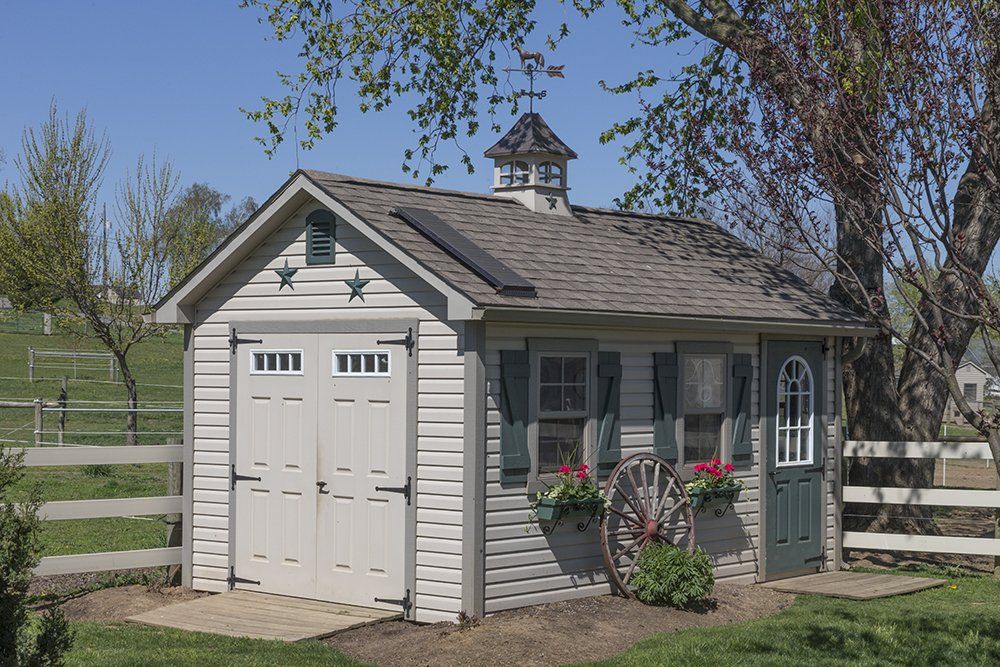 Small shed with a gray exterior, green door, and flower boxes; set in a grassy yard, under a blue sky.