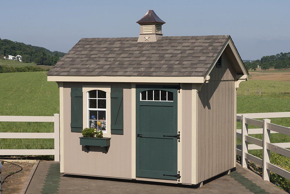 Tan shed with green door and shutters, small window, brown roof, and copper-colored decorative top, outside.