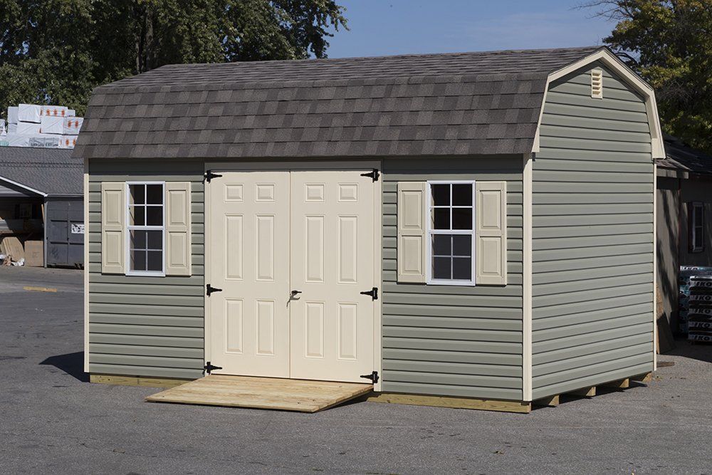 Green shed with barn-style roof, beige doors, and shutters. A wooden ramp leads to the entrance.