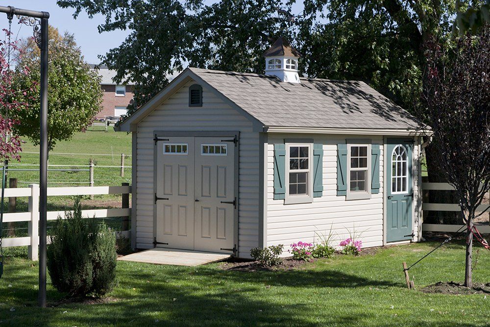 Light beige shed with double doors, two windows with green shutters, and a small cupola on a grassy lawn.