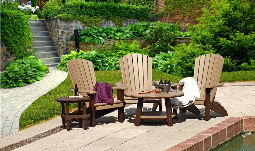 Outdoor seating area with three brown Adirondack chairs, a table, and lush greenery.