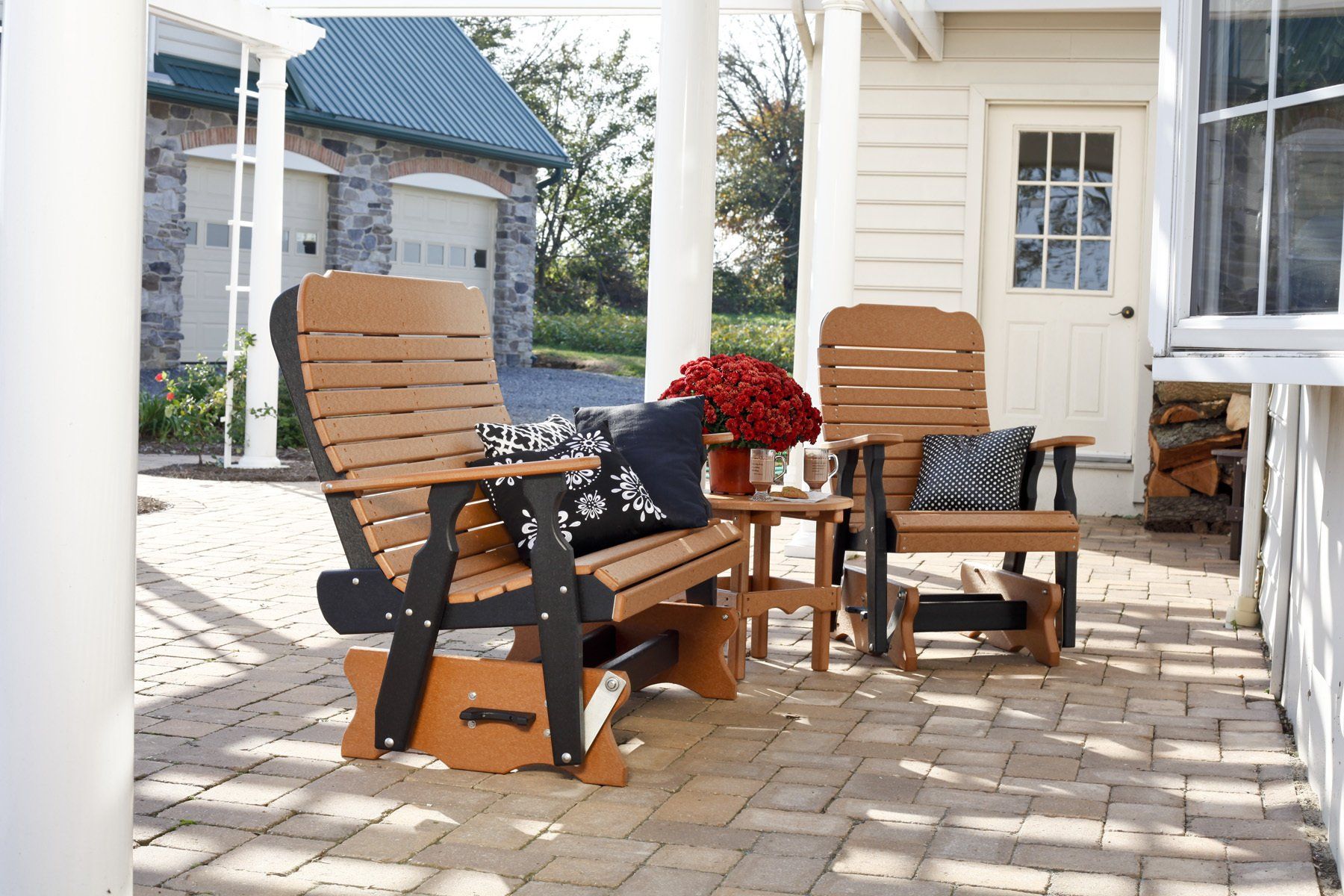 Two wooden gliding chairs and small table on a patio, under a pergola, near a house with stone accents.