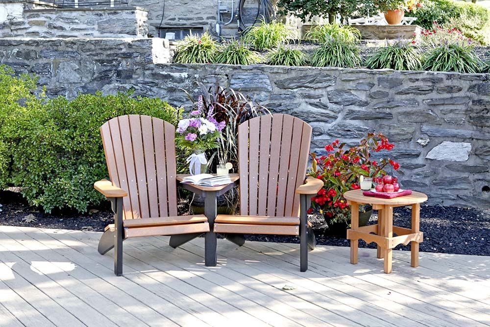 Two brown Adirondack chairs with a small table on a patio with a stone wall backdrop.