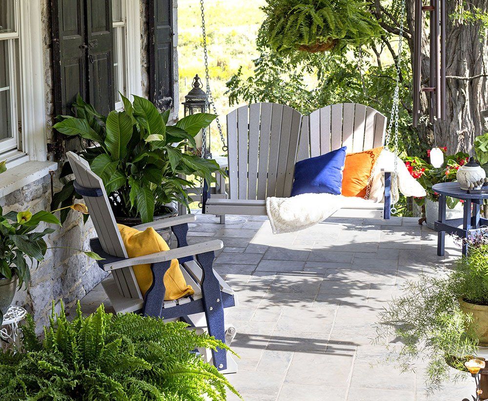 Porch with wooden swing, chair, and plants; stone wall, yellow and blue accents; sunny outdoor setting.