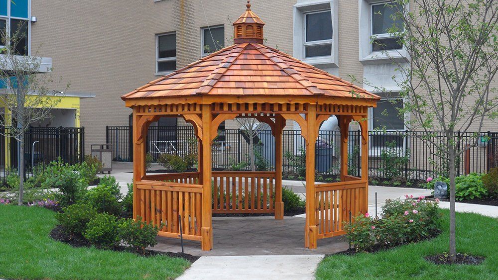 Wooden gazebo in a green grassy area with a building in the background.