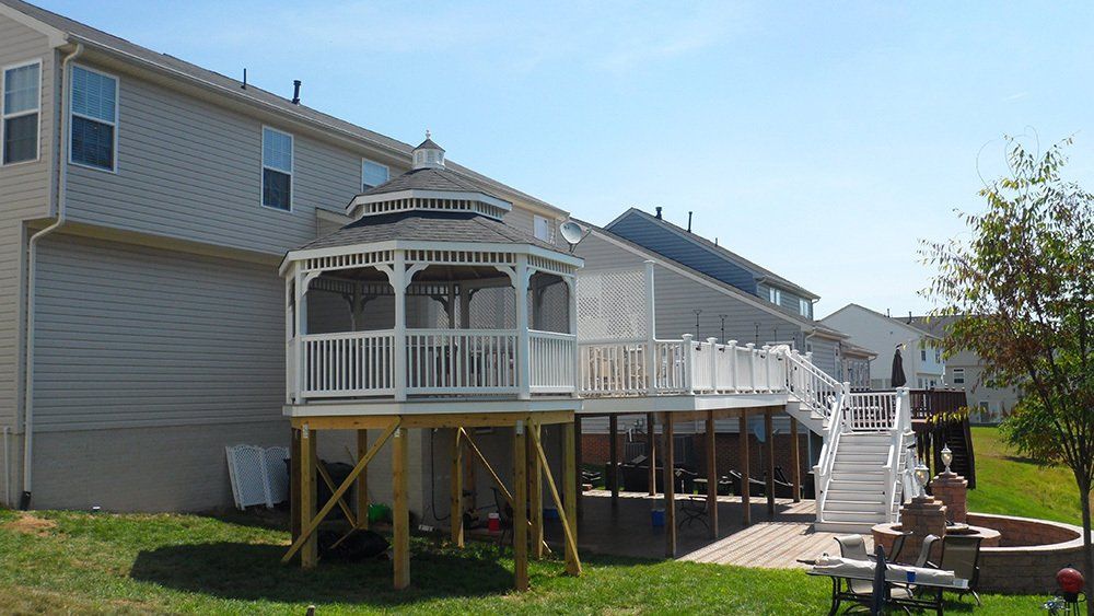 White deck and gazebo attached to a beige house, supported by wooden posts. Sunny day.