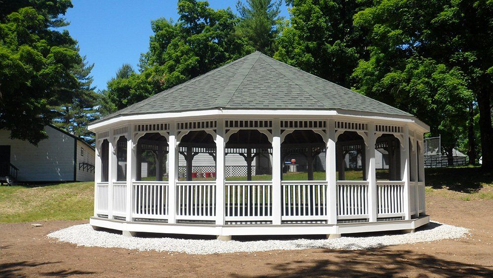 White gazebo with green roof in a park, surrounded by trees and gravel.