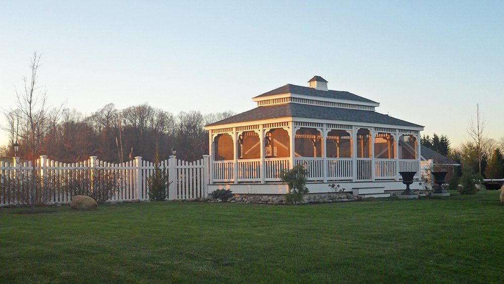 White gazebo on a grassy lawn, surrounded by a white picket fence, trees in the background, daytime.