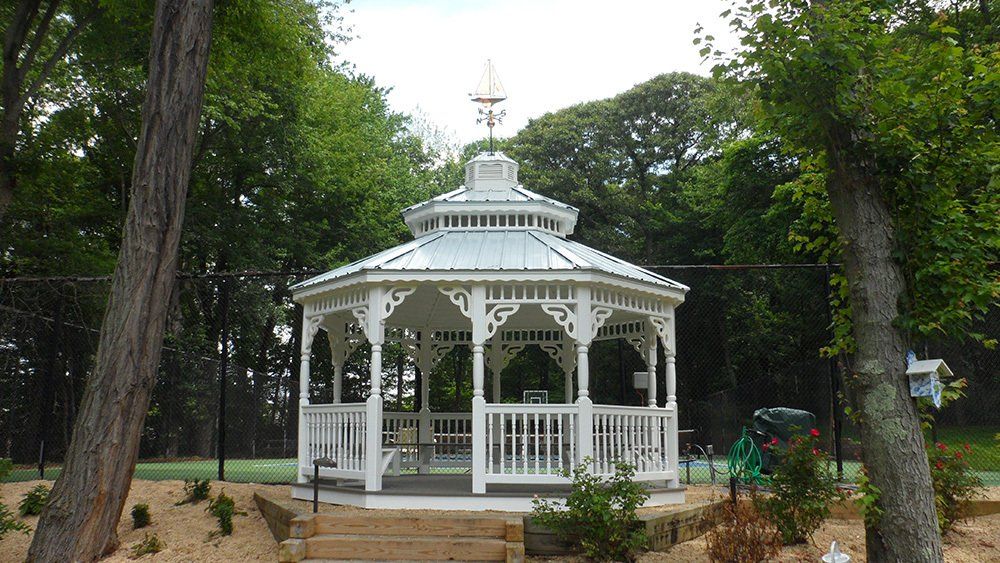 White gazebo with decorative trim and weather vane, set amidst trees and landscaping.