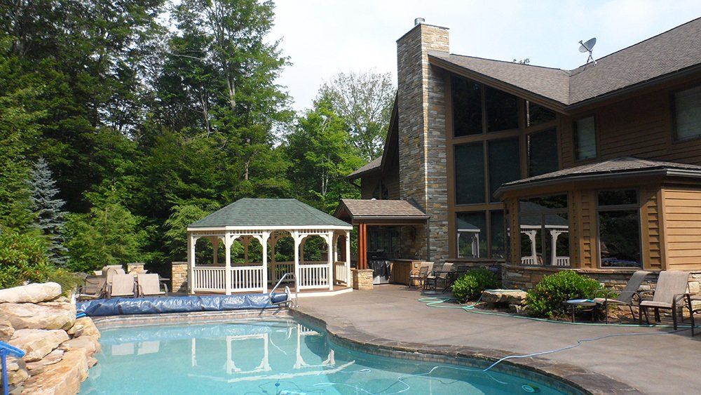 Backyard with pool, gazebo, and large house with stone chimney and wood siding; trees in background.