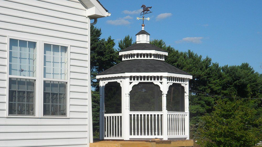White gazebo with black roof and horse weathervane, next to a white house with window.