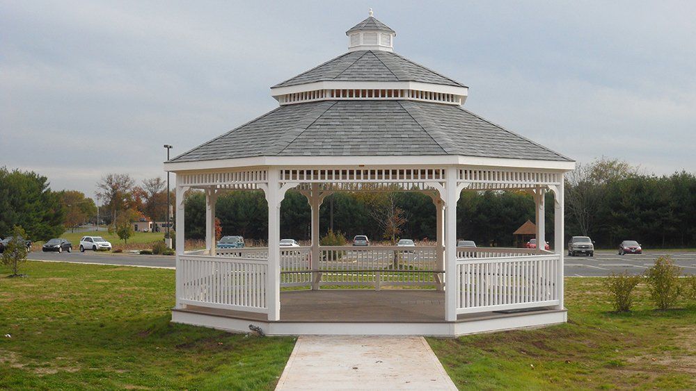 White gazebo with gray roof on a green lawn, sidewalk in front, vehicles visible in background.