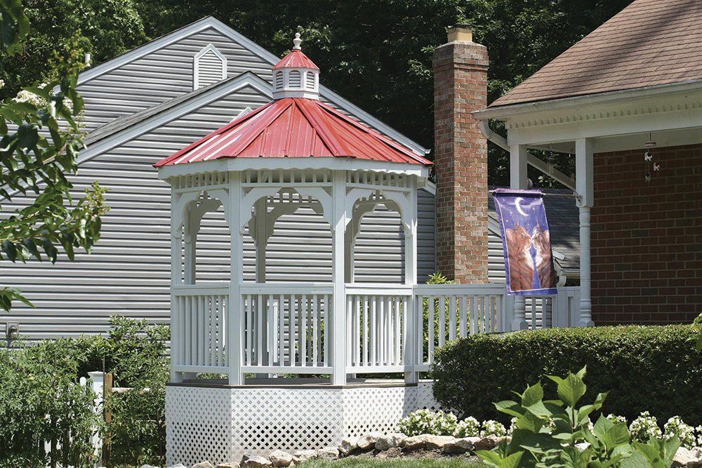 White gazebo with red roof in a yard, beside a house with a brick chimney and a blue and purple banner.