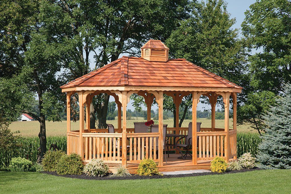 Wooden gazebo with brown roof in a green yard, trees in the background.