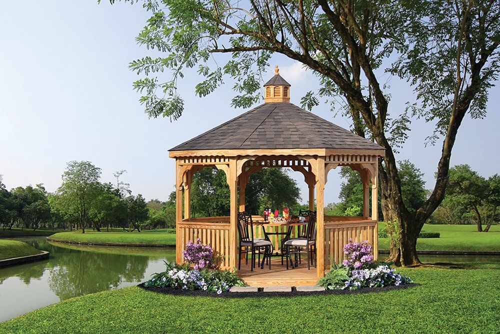 Wooden gazebo with a table and chairs beside a lake, under a tree.
