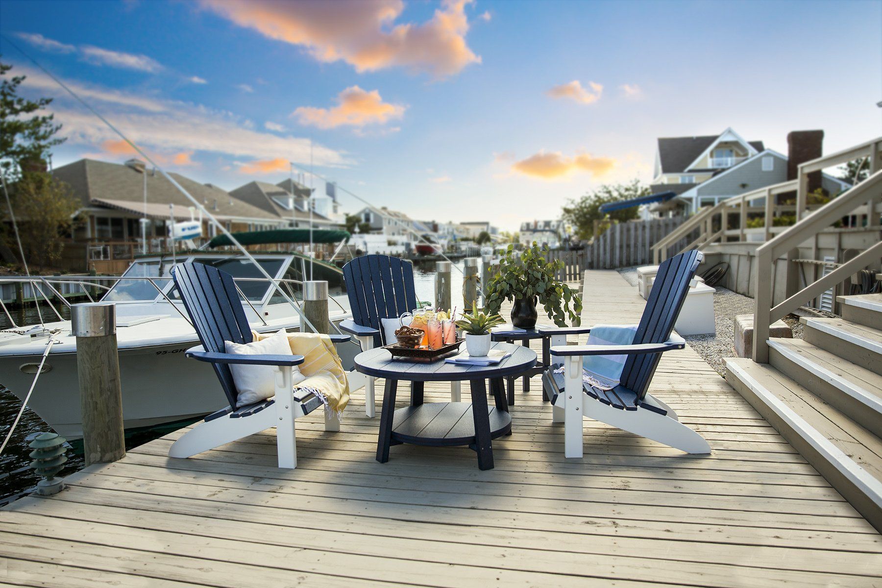 Wooden dock with Adirondack chairs and table; boats and houses in background.