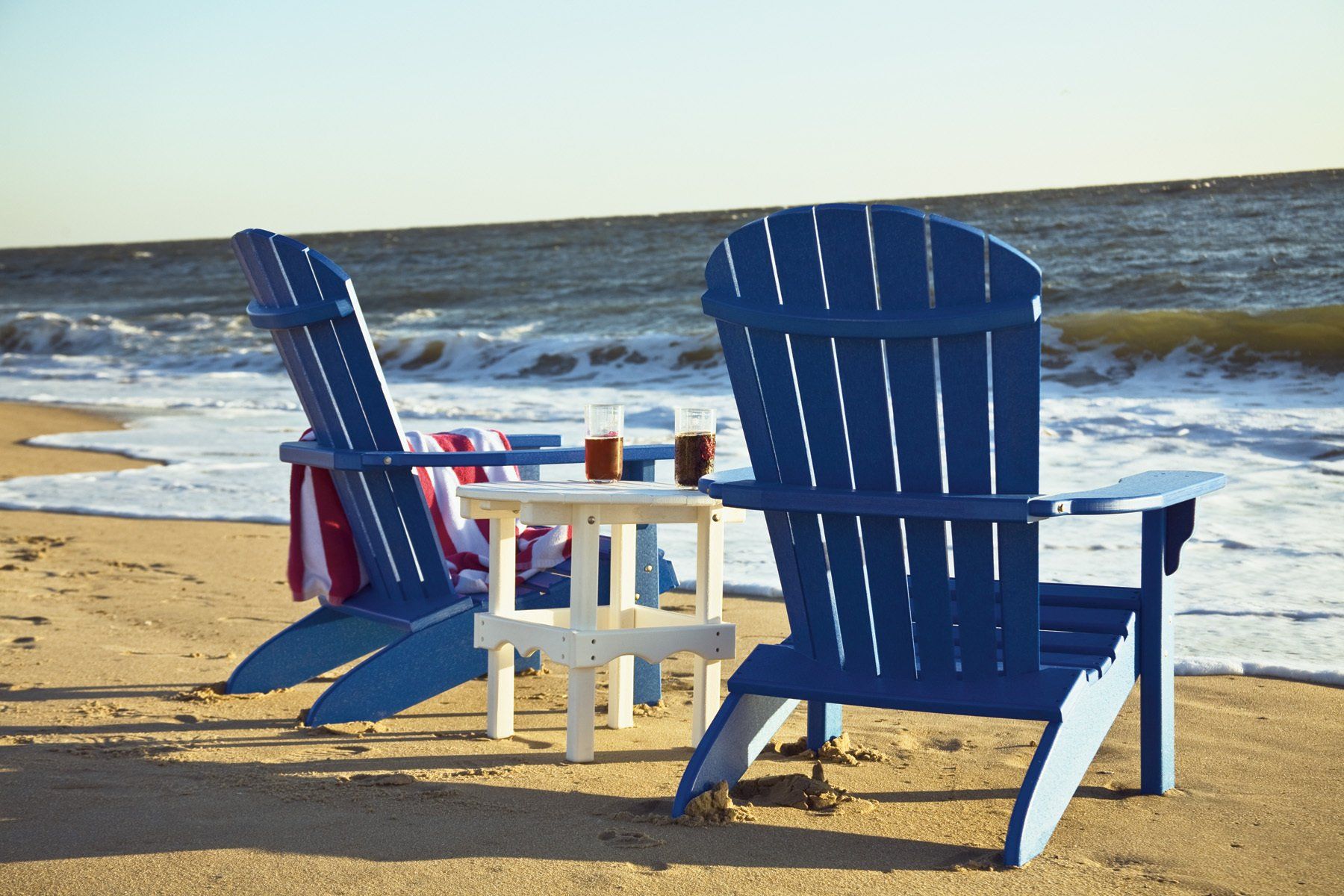 Two blue Adirondack chairs face the ocean, with a small table between them holding two drinks. A towel hangs on one chair.