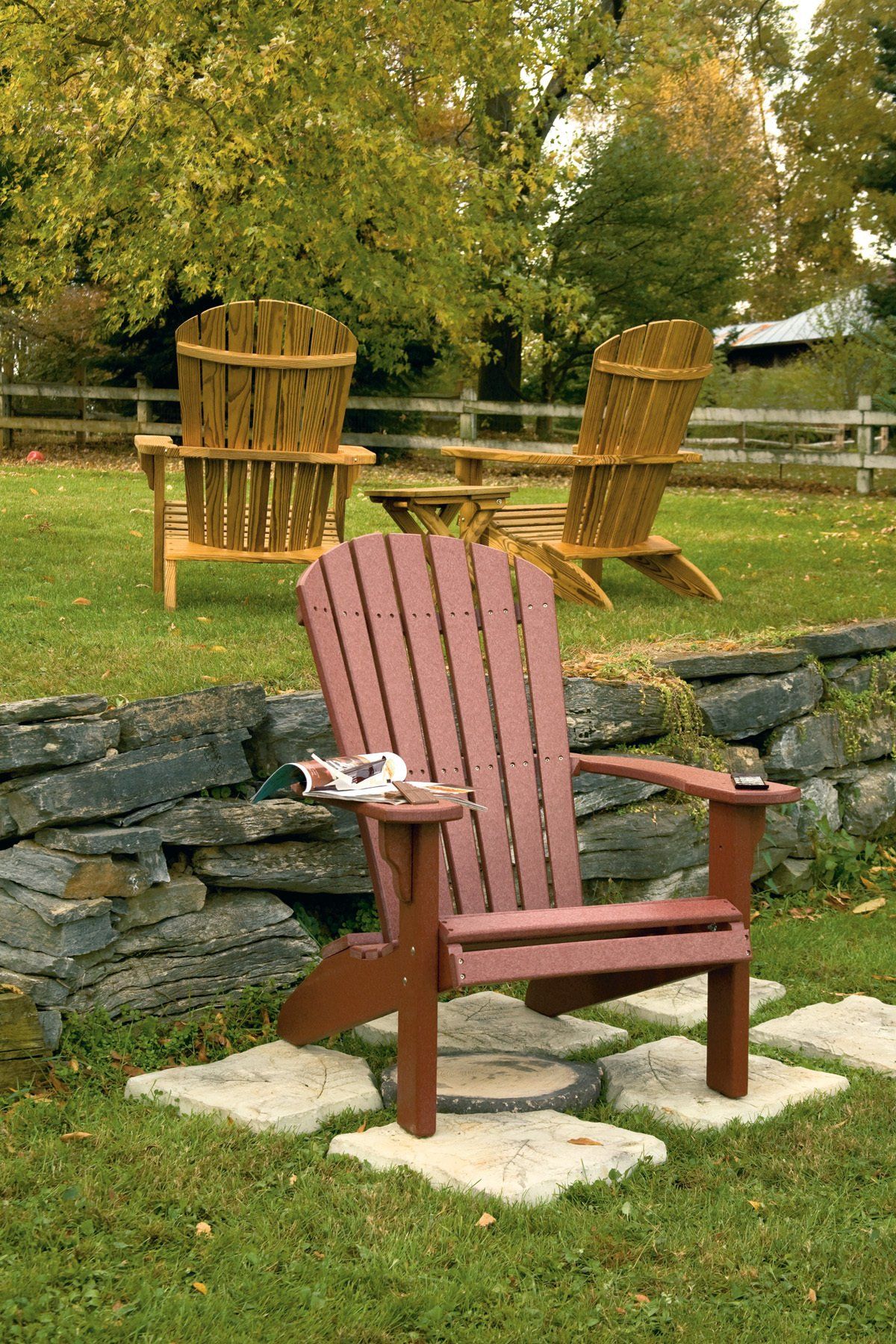 Red Adirondack chair on stone pavers, two rustic wood chairs, and a table in a grassy yard by a stone wall.