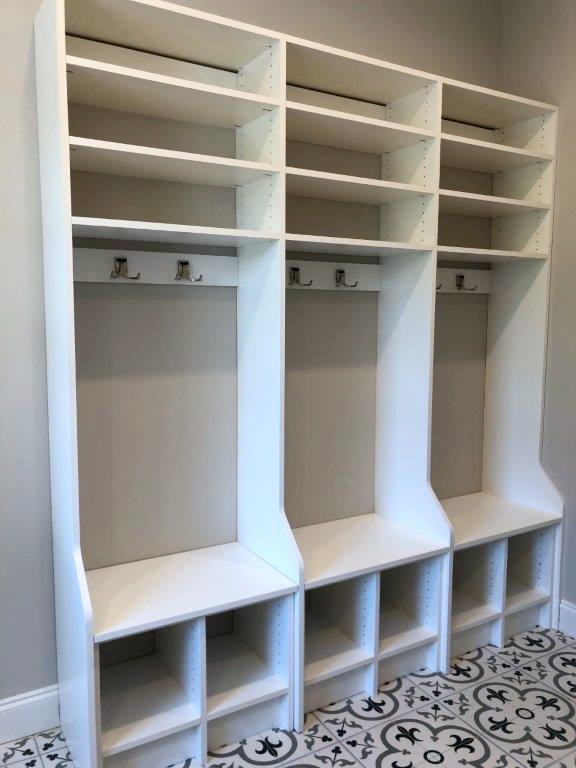 Three white, floor-to-ceiling wooden storage cubbies with bench seating, coat hooks, and shelves against a gray wall.