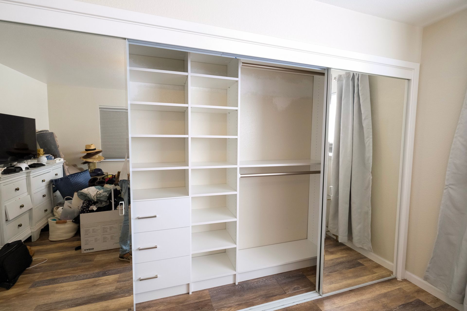 A reach-in closet with white shelving, three drawers, and a hanging rod, flanked by mirrored sliding doors in a room.