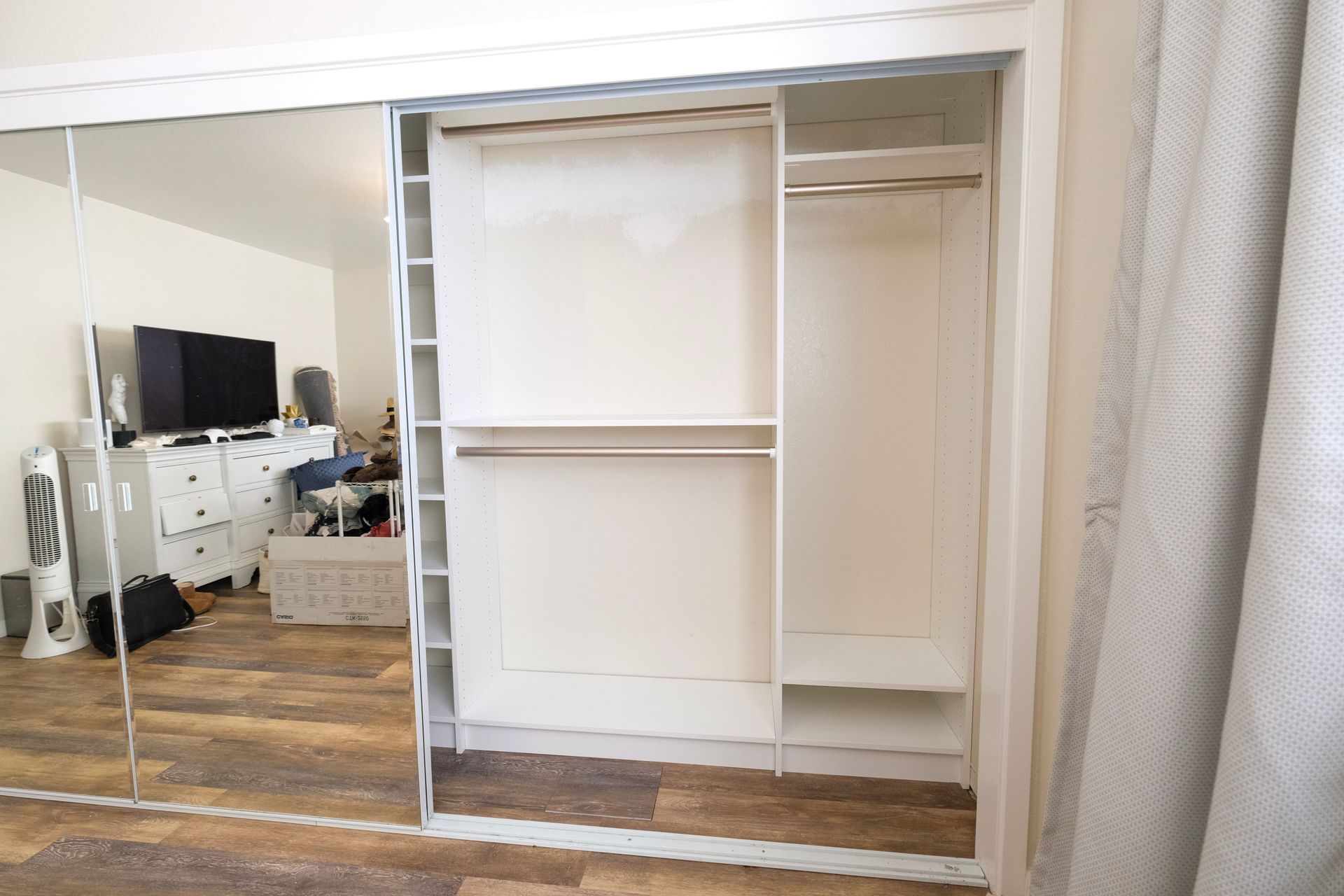 An open white reach-in closet with shelves and hanging rods, seen through partially opened sliding mirrored doors.