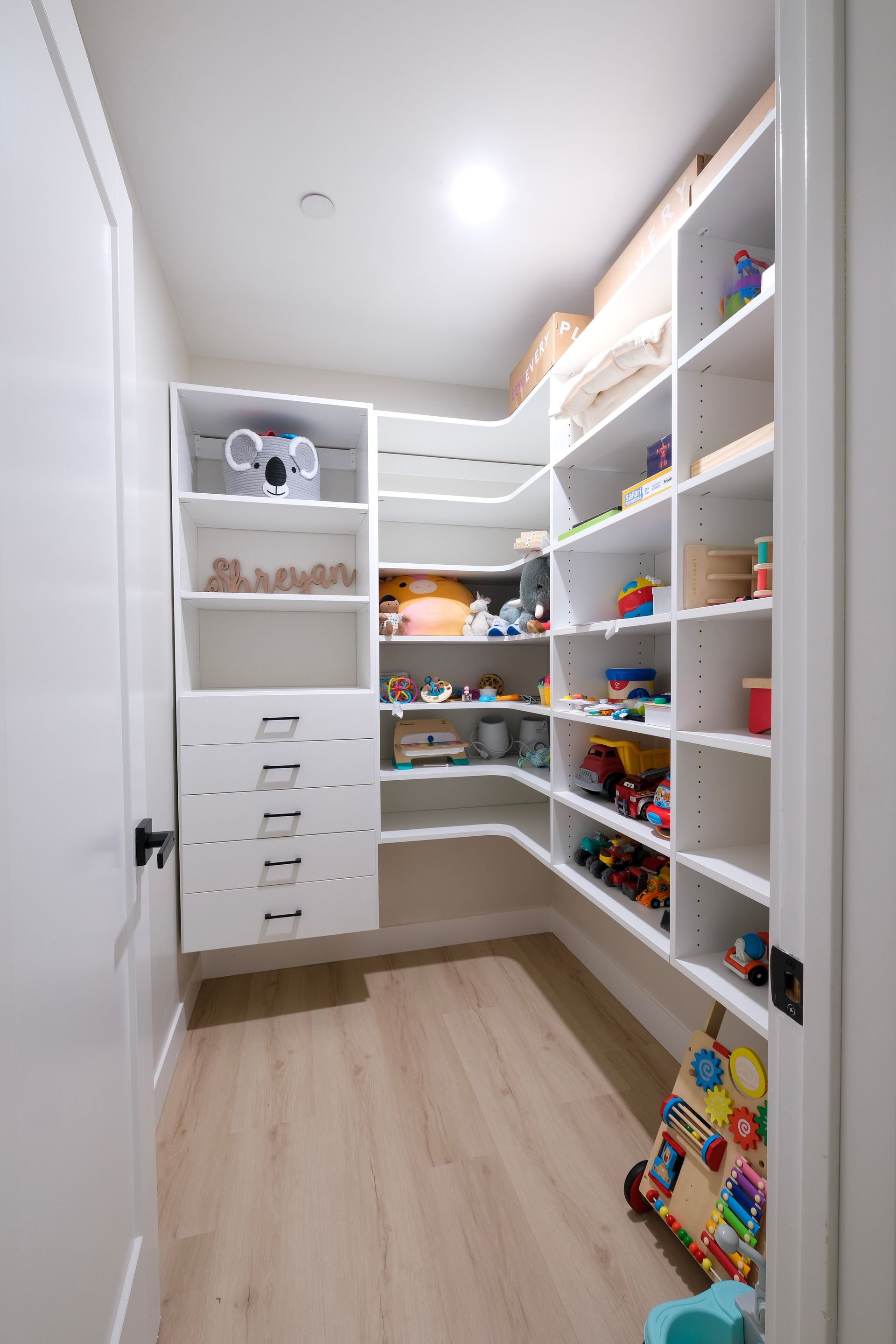 A white, L-shaped walk-in closet with shelves and drawers, organized with children's toys and books.