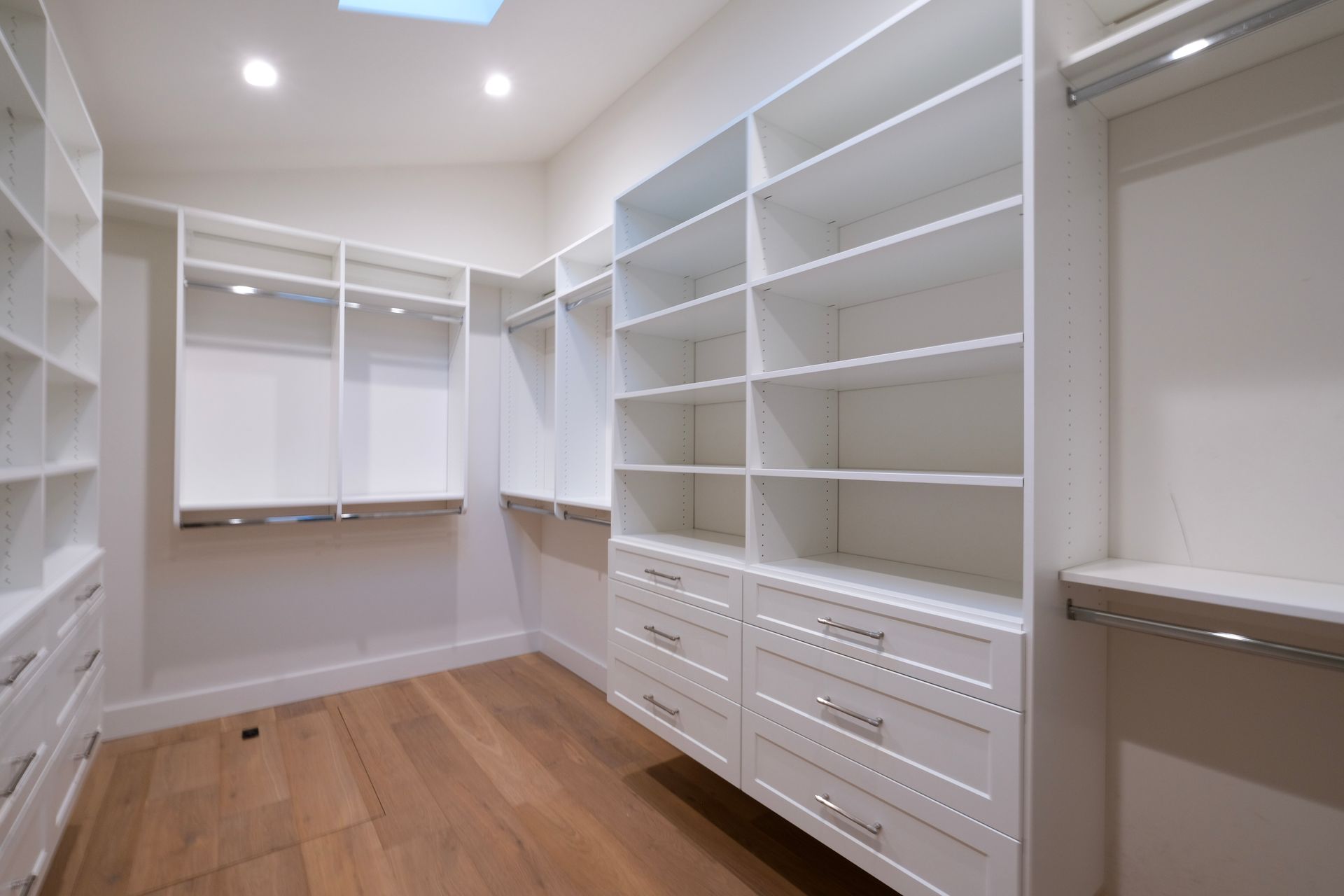 A brightly lit, modern walk-in closet with white custom shelving, hanging rods, and drawers on a hardwood floor.