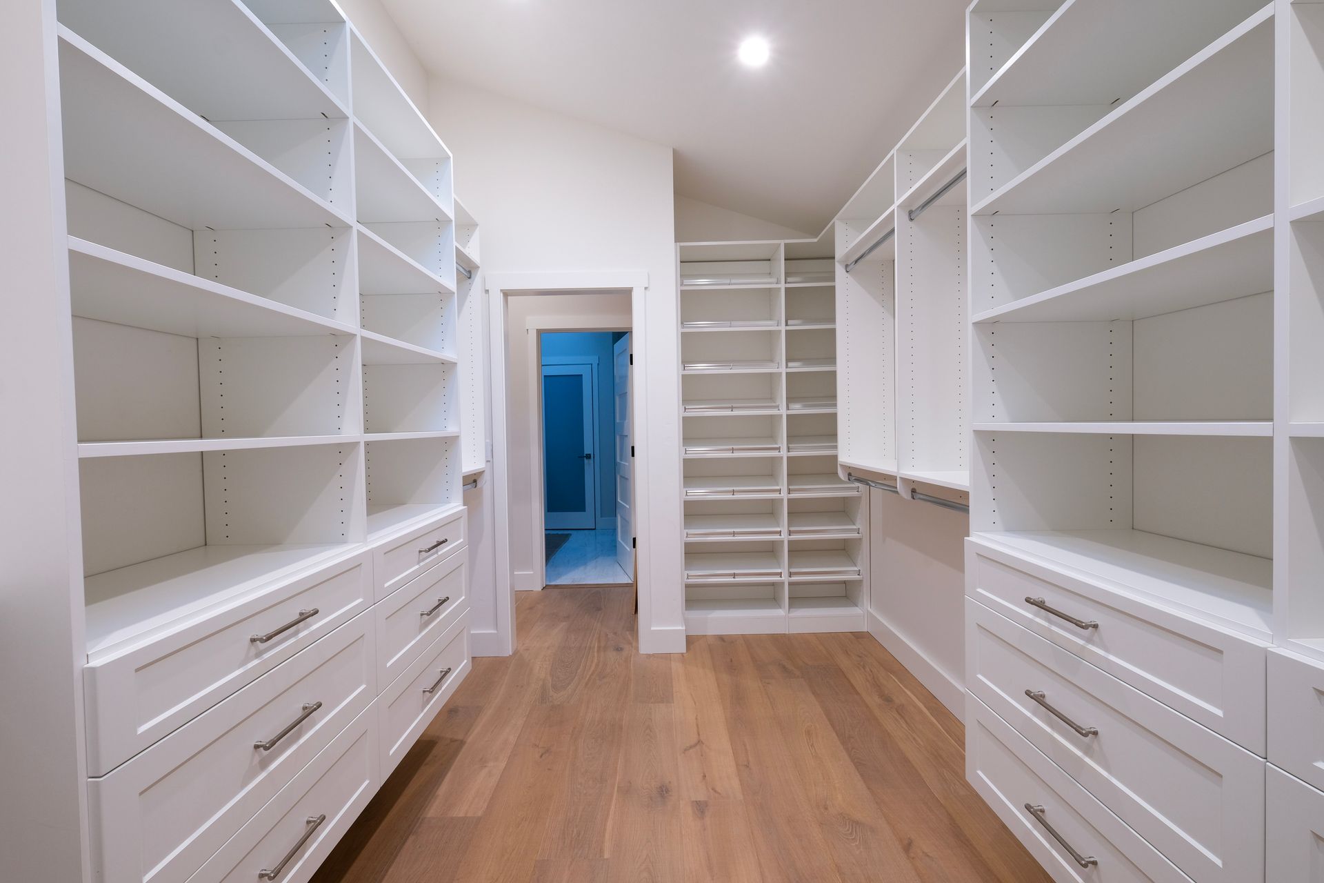A bright, walk-in closet with white shelving, stacked drawers, and light wood flooring, leading to a doorway.